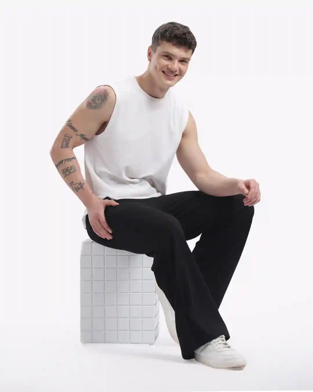 Man sitting on a white cube wearing a white tank top and black pants against a white background