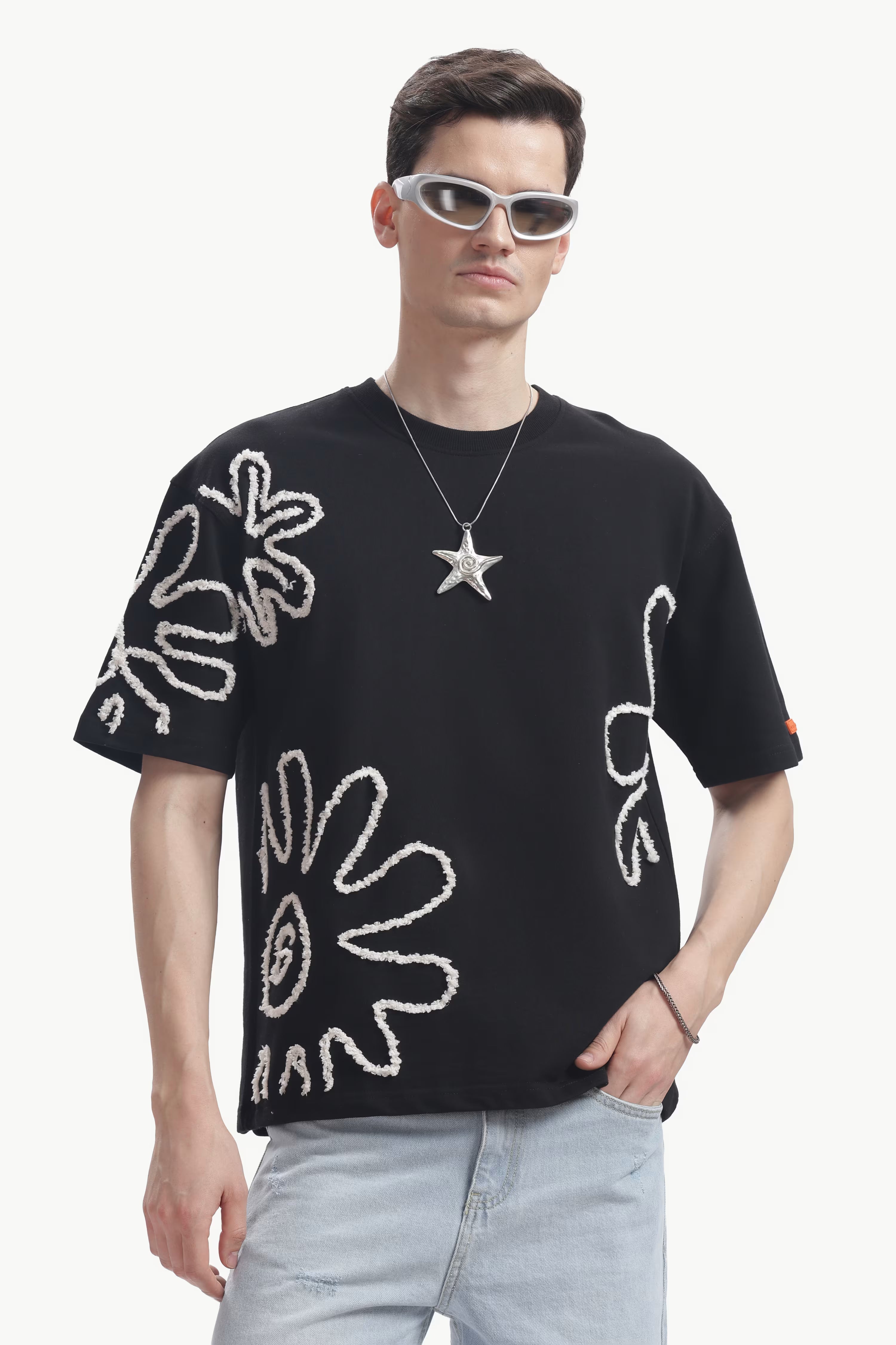 Man wearing a black t-shirt with white floral designs, light blue jeans, and sunglasses on a white background