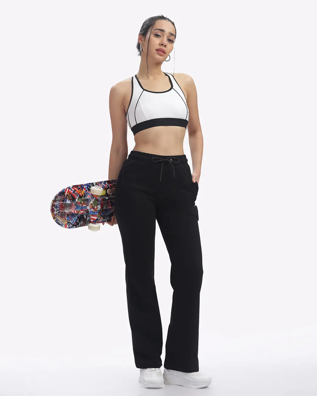 Woman holding a skateboard wearing a sports top and black joggers on a white background