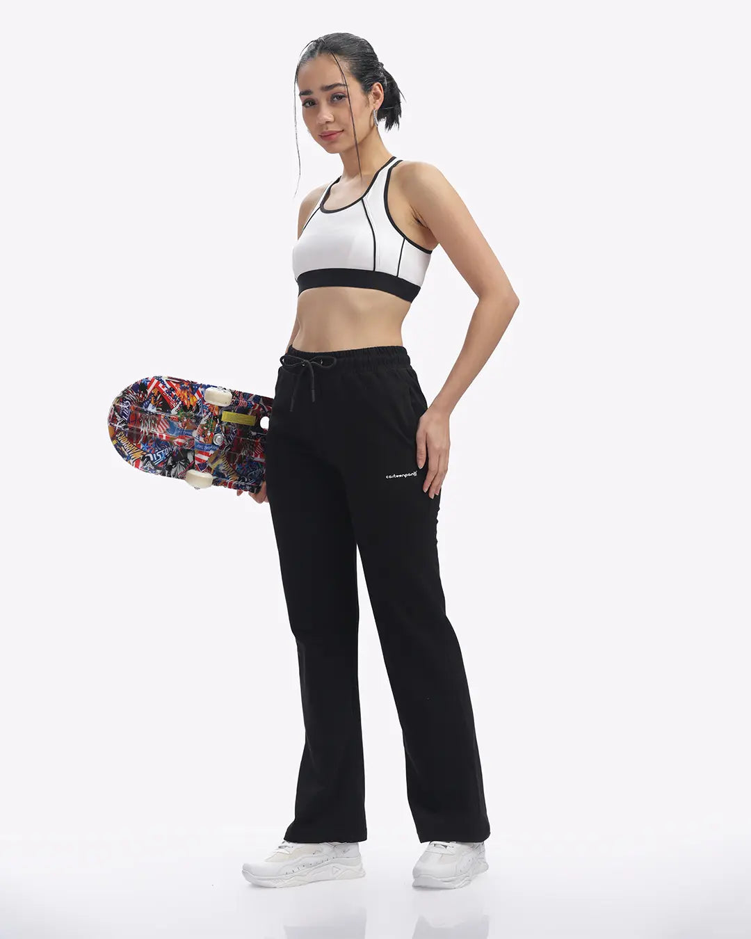 Woman holding a colorful skateboard wearing a white sports bra and black pants on a white background